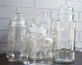 May include: Four clear glass jars with lids. The jars are different sizes and shapes. The jars are on a dark brown surface.
