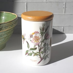 May include: A white ceramic canister with a wooden lid. The canister is decorated with a botanical illustration of a dog rose and a bee. The text "Rosa Canina Dog Rose" is printed on the canister.