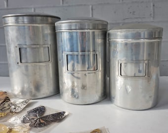 May include: Three silver metal canisters with labels. The canisters are all the same size and shape, and they have a slightly distressed look. They are sitting on a white surface.