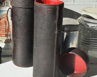 May include: Two brown leather cylindrical cases with red lining. The cases are closed and sitting on a white surface. One case has a lid that is open and is sitting on the surface next to the cases.