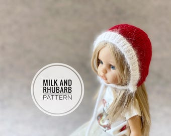Könnte beinhalten: Eine Puppe trägt eine rot-weiße Strickmütze mit weißem Rand und weißer Schleife. Die Mütze trägt ein Etikett mit der Aufschrift "Milk and Rhubarb Pattern".