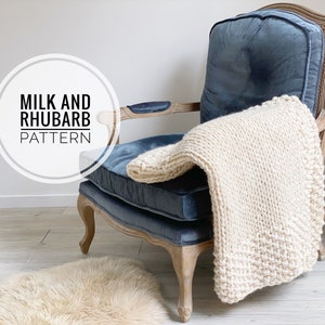 May include: A white knitted throw blanket draped over a blue velvet armchair with wooden arms and legs. The chair is in front of a white wall and a light wood floor. The text "Milk and Rhubarb Pattern" is in a white circle on the left side of the image.