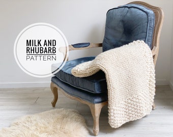 May include: A cream-colored knitted blanket draped over a blue velvet armchair. The chair has a wooden frame and a sheepskin rug is on the floor. The text "MILK AND RHUBARB PATTERN" is in a circle.