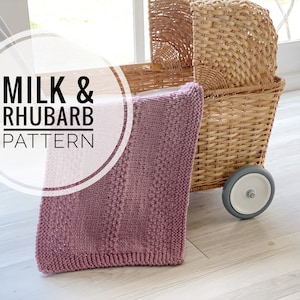 May include: A pink knitted baby blanket with the text "Milk & Rhubarb Pattern" in a white circle, resting on a wicker doll stroller with a gray wheel.