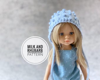 May include: A light blue knitted doll hat with a bobble detail and long ties. The hat is on a doll with blonde hair and a blue polka dot dress. The text "Milk and Rhubarb Pattern" is on a white circle.