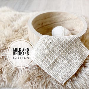 May include: A square, cream-coloured knitted item with a textured pattern, resting on a fluffy, light brown surface. A wooden bowl and a ball of white yarn are in the background. The text "MILK AND RHUBARB PATTERN" is in a circle.