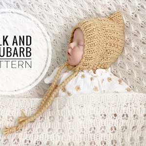 Può includere: Un bambino che indossa un cappello di lana beige con un lungo laccio, sdraiato su una coperta di uncinetto bianca. Il testo "Milk and Rhubarb Pattern" è visibile nell'immagine.