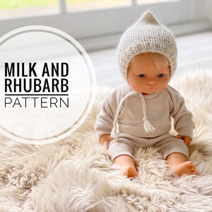 May include: A doll dressed in a beige knit hat, top, and trousers, with a drawstring. The doll is seated on a fluffy, cream-coloured rug. The image includes the text "MILK AND RHUBARB PATTERN" in a circular design.