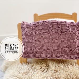 May include: A knitted blanket in a mauve color with a textured, woven pattern. The blanket is draped over a small wooden chair. The text "MILK AND RHUBARB PATTERN" is in a white circle in the lower left corner.