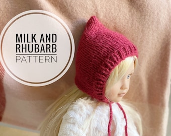 Könnte beinhalten: Nahaufnahme einer roten gestrickten Puppenmütze mit spitzer Spitze und einer Schleife. Die Mütze wird von einer Puppe mit blonden Haaren getragen. Der Text "MILK AND RHUBARB PATTERN" ist in einem weißen Kreis sichtbar.