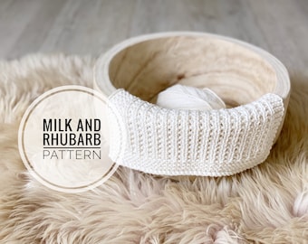 May include: White knitted baby blanket in a wooden bowl on a fluffy beige rug. The text "Milk and Rhubarb Pattern" is in a circle on the rug.