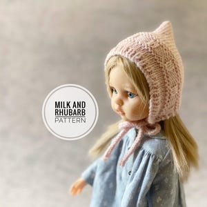 May include: A doll wearing a light pink knitted bonnet with a pointed top and a matching tie. The doll has blonde hair and is wearing a blue dress. The image includes a circular logo with the text "MILK AND RHUBARB PATTERN".