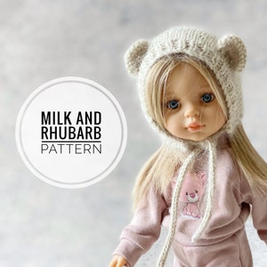 May include: A knitted doll hat with bear ears in a cream colour. The hat has a long tie that hangs down the front of the doll. The doll is wearing a pink sweatshirt with a small bear graphic and pink trousers. The text "Milk and Rhubarb Pattern" is visible in the image.