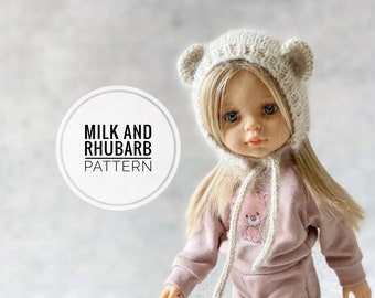 May include: A knitted doll hat with bear ears in a cream color. The hat has a long tie that hangs down the front of the doll. The doll is wearing a pink sweatshirt with a small bear graphic and pink pants. The text "Milk and Rhubarb Pattern" is visible in the image.