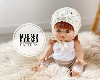 Könnte beinhalten: Eine Puppe trägt eine weiße Häkelmütze mit einem strukturierten Muster und ein weißes Tanktop mit einer beige Hose. Die Puppe sitzt auf einer weißen, flauschigen Oberfläche. Der Text "Milk and Rhubarb Pattern" ist in einem weißen Kreis sichtbar.