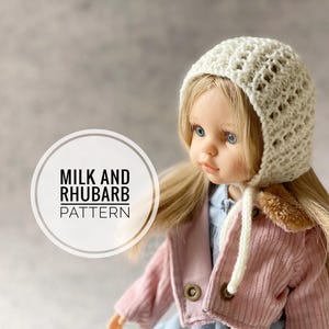 May include: A doll wearing a cream-colored crocheted bonnet with a textured pattern. The doll has blonde hair, blue eyes, and is dressed in a pink corduroy jacket. The image includes the text "MILK AND RHUBARB PATTERN" in a circular design.
