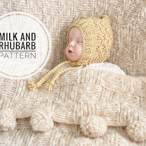 Puede incluir: Un bebé con un gorro de punto beige con un pompón en la parte superior, acostado sobre una manta de punto beige con pompones. La manta tiene un patrón tejido. El texto "Milk and Rhubarb Pattern" es visible en la imagen.