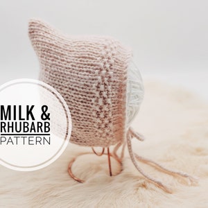 Könnte beinhalten: Hellrosa gestrickte Babyhaube mit spitzer Spitze, auf einem Drahtständer präsentiert. Die Haube hat ein strukturiertes Strickmuster und Bindebänder. Der Text "MILK & RHUBARB PATTERN" befindet sich in einem weißen Kreis.