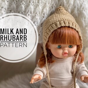 Pode incluir: Uma boneca usando um chapéu de tricô bege com a parte superior pontiaguda e um suéter de tricô bege. A boneca tem cabelo ruivo e olhos azuis. O texto "MILK AND RHUBARB PATTERN" é visível na imagem.