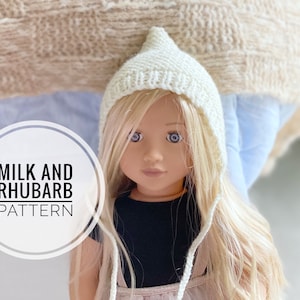 Może przedstawiać: Biały, dzianinowy kapelusz dla lalki z ostrym kapturem i długimi sznurkami. Kapelusz nosi lalka o blond włosach i niebieskich oczach. Na zdjęciu widoczny jest tekst "MILK AND RHUBARB PATTERN".