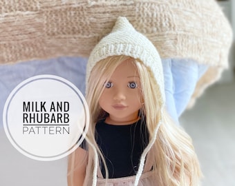 Könnte beinhalten: Eine weiße gestrickte Puppenmütze mit einer spitzen Kapuze und langen Bändern. Die Mütze wird von einer Puppe mit blonden Haaren und blauen Augen getragen. Der Text "MILK AND RHUBARB PATTERN" ist auf dem Bild zu sehen.