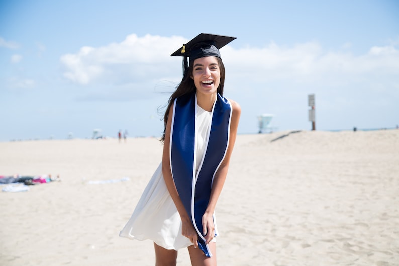 May include: A woman wearing a white dress and a blue graduation sash smiles on a sandy beach. She is wearing a black graduation cap.