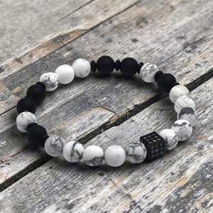 Peut inclure: Un bracelet de perles composé d'une alternance de perles sphériques noir mat et de perles blanches marbrées. Le design comprend une perle carrée noire unique avec une surface texturée et cloutée, intégrée à la disposition circulaire.