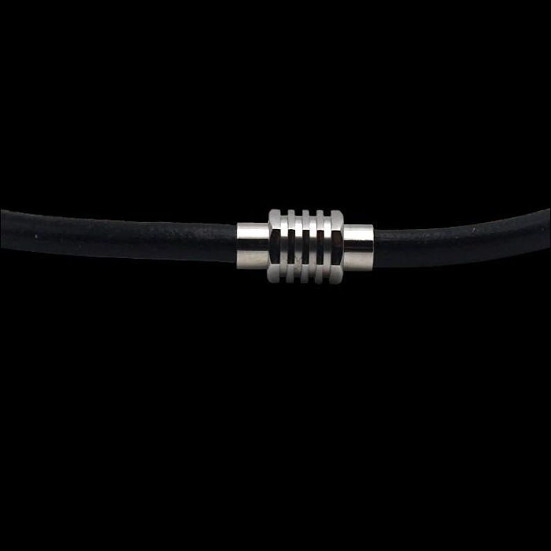 Puede incluir: Primer plano de un collar negro con un cierre cil&iacute;ndrico plateado. El cierre tiene un dise&ntilde;o acanalado y est&aacute; centrado en el collar. El fondo es negro s&oacute;lido.