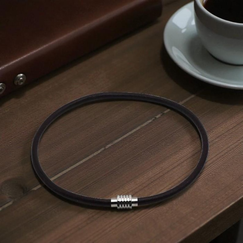 Puede incluir: Un collar negro con un cierre plateado descansa sobre una superficie de madera. El collar es circular y parece estar hecho de un material flexible. Un cuaderno encuadernado en cuero marr&oacute;n y una taza y platillo blancos est&aacute;n en el fondo.