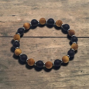 May include: A circular beaded bracelet featuring alternating matte black spherical beads and light brown wood-grain beads, separated by small, polished silver-toned metal spacer beads arranged in a repeating pattern around the entire circumference.