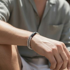Peut inclure: Un bracelet en cuir blanc tressé avec un fermoir en métal noir est porté au poignet. Le bracelet est composé de deux brins de cuir tressé. La personne porte une chemise vert clair.