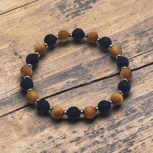 May include: A beaded bracelet featuring alternating round black porous lava stone beads and smooth light brown wooden beads. Small, polished silver-toned metal spacer beads are placed between each of the larger stone and wooden beads.