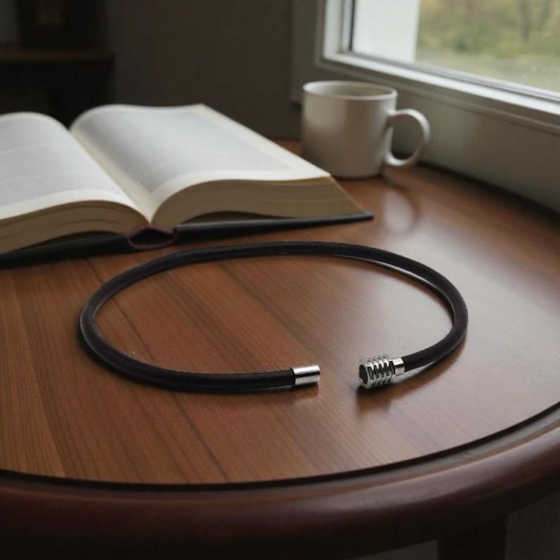Puede incluir: Un collar de cuero negro con un cierre plateado descansa sobre una mesa de madera. El collar est&aacute; abierto, revelando el mecanismo de cierre. Un libro abierto y una taza blanca est&aacute;n en el fondo.