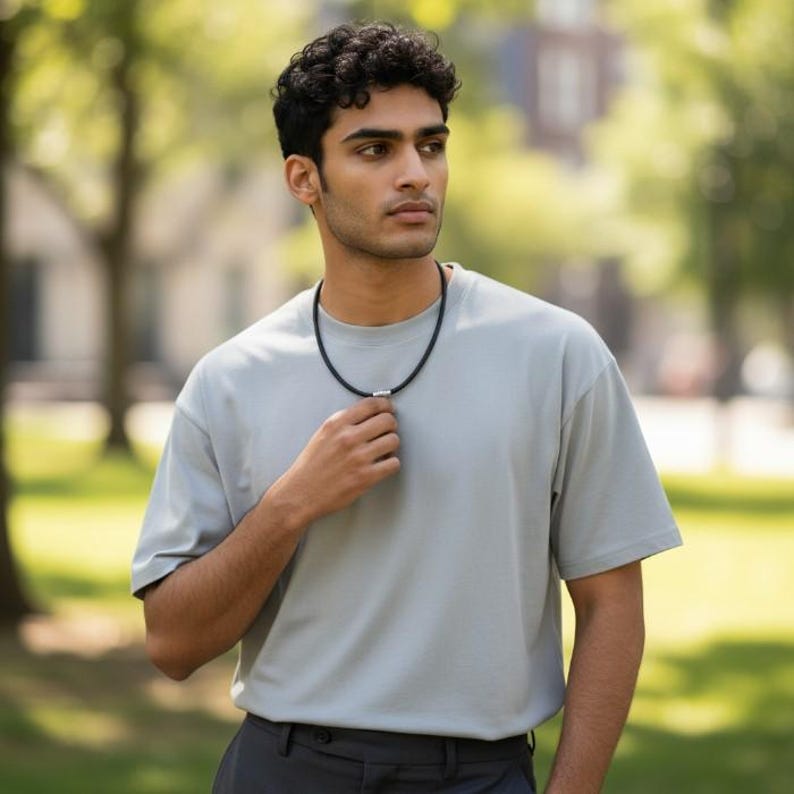 Puede incluir: Un collar negro con un colgante cil&iacute;ndrico de color plateado. El collar lo lleva una persona que viste una camiseta gris claro. El fondo es una escena borrosa al aire libre con &aacute;rboles y follaje verdes.