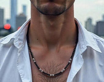 May include: A black beaded necklace with silver accents. The necklace is worn over a white button-down shirt. The background features a blurred cityscape with a cloudy sky.