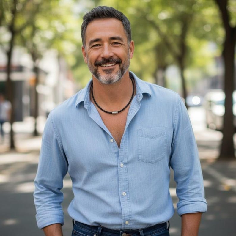 Puede incluir: Un hombre con una camisa abotonada azul claro y vaqueros azul oscuro. Lleva un collar negro con un cierre plateado. El fondo es una escena callejera borrosa con &aacute;rboles.