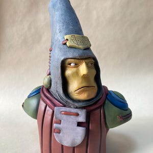 May include: A ceramic bust of a fictional character wearing a blue pointed hat, green and blue armour, and a red and grey chest piece. The character has a yellow face with a serious expression and a small, gold plaque on the hat that reads "The F".