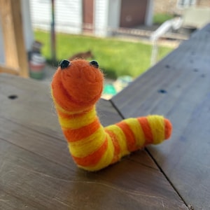May include: A handmade felt worm sculpture. The worm is orange and yellow, with black bead eyes. The worm's body is segmented with alternating bands of orange and yellow. The worm is posed on a wooden surface.