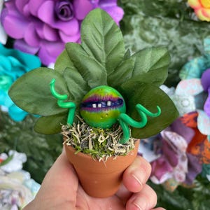 May include: A handcrafted sculpture of a carnivorous plant in a small terracotta pot. The plant features a green, spotted head with a purple mouth and white teeth, surrounded by green tendrils and large green leaves. The pot is filled with moss.