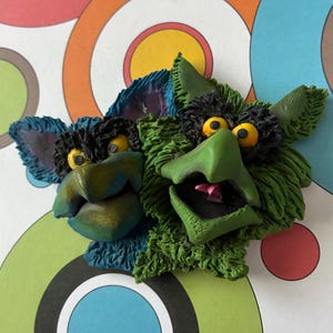 May include: Two whimsical, handmade clay sculptures of fantastical creatures. One is blue, the other green, both with yellow eyes, black hair, and open mouths. The background features a colorful, retro-style pattern of overlapping circles.
