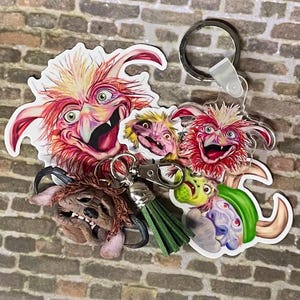 May include: A collection of colorful, whimsical keychains featuring fantasy creature designs. The keychains include various characters with vibrant red, yellow, green, and blue hues, attached to metal keyrings with green tassels. The designs are detailed and eye-catching.