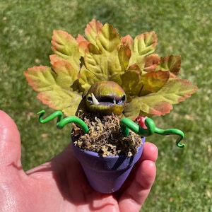 May include: A quirky, handmade plant sculpture in a small purple pot. The plant features large, textured green and orange leaves, a toothy mouth, and green, curling vines. The sculpture is set against a blurred green background.