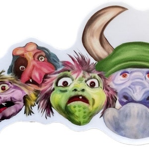 May include: A close-up of four colourful, furry monster puppets with large eyes. The puppets are arranged in a row, with the first puppet on the left having purple fur and yellow hair, the second puppet having pink skin and blue hair, the third puppet having green skin and pink hair, and the fourth puppet having blue skin and green horns.