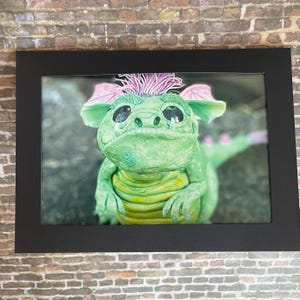 May include: A framed image of a green dragon sculpture with pink and purple accents. The dragon has large eyes, a friendly expression, and a yellow belly. The artwork is set against a blurred background and framed in a black border.