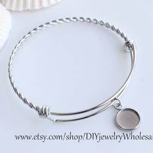 May include: A silver bangle bracelet with a twisted wire design and a round, blank charm pendant. The bracelet is open and adjustable.