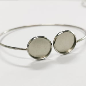 May include: Silver bangle bracelet with two round, silver-toned, bezel settings. The settings are empty and ready to be filled with resin or other materials.