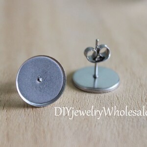 May include: Two silver earring blanks with a round base and a post for attaching earrings. The blanks are on a wooden surface.