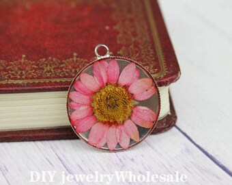 Op de afbeelding: Een ronde, zilverkleurige hanger met een geperste roze bloem erin. De bloem heeft een gele kern en is omgeven door een heldere hars.