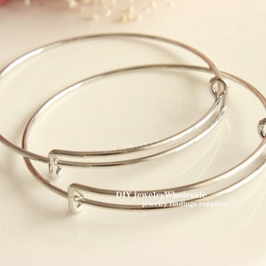 May include: Two silver bangle bracelets, one slightly larger than the other, are stacked on top of each other. The bracelets are made of thin metal and have a simple, elegant design.