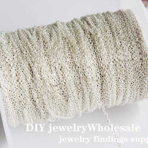 Puede incluir: Un carrete de cadena plateada, enrollado alrededor de un carrete de plástico blanco. La cadena es fina y delicada, y el carrete está etiquetado con "DIY jewelryWholesale jewelry findings supplies".